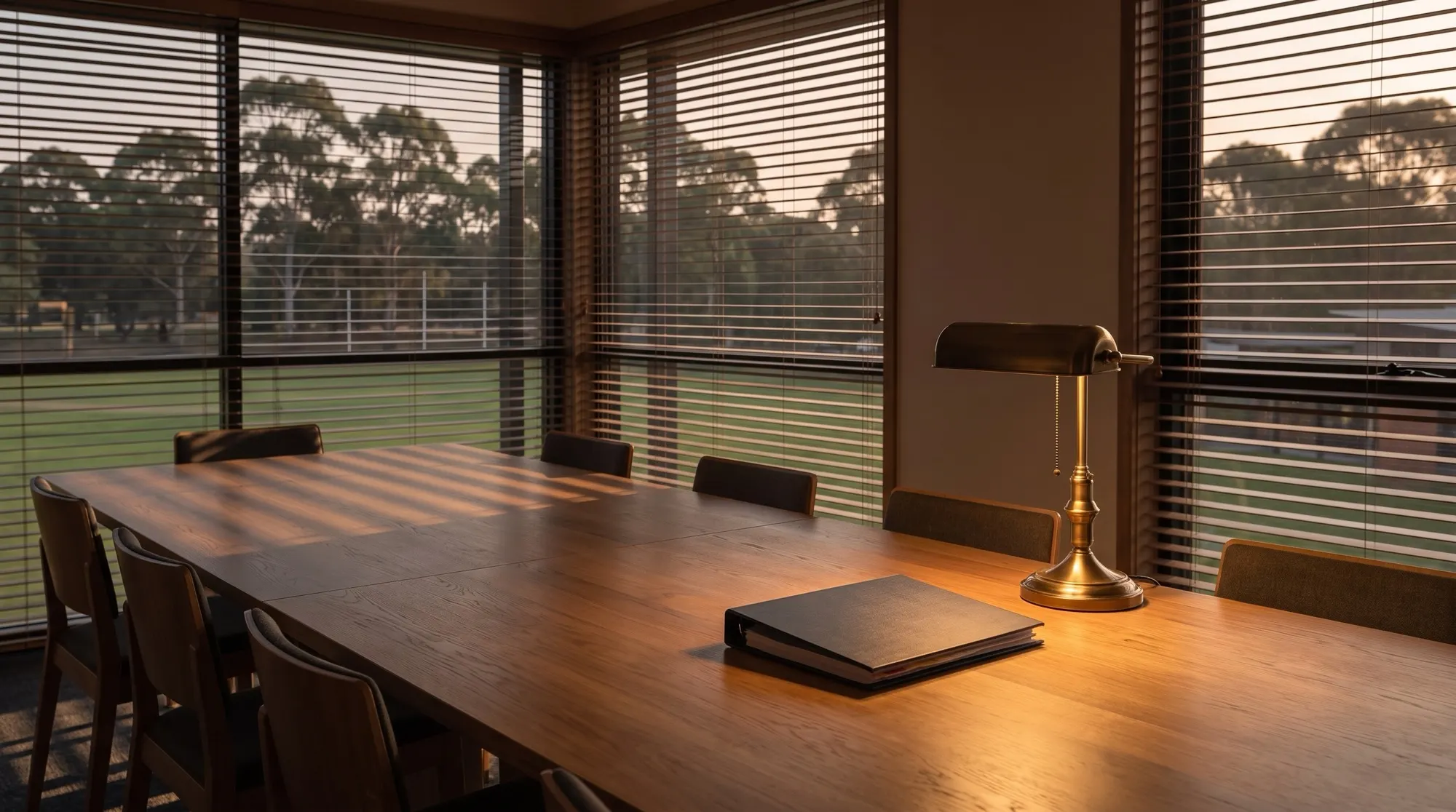 An empty school boardroom at dusk, chairs pushed in, a single closed binder and a reading lamp left on, warm light falling through timber blinds across the table.